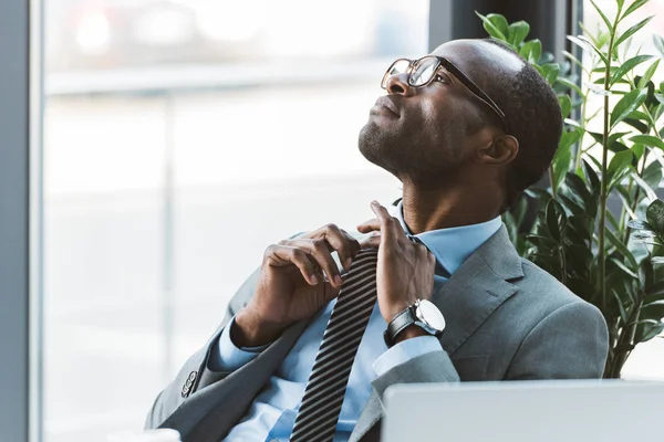 Young african american businessman in eyeglasses adjusting necktie and looking away in office — Stock Photo