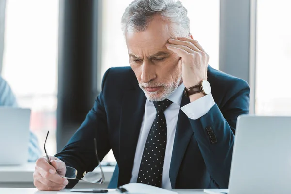 Tired senior businessman with headache holding eyeglasses and looking down at workplace — Stock Photo