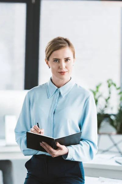 Schöne Geschäftsfrau, die Notizen macht und in die Kamera schaut — Stockfoto