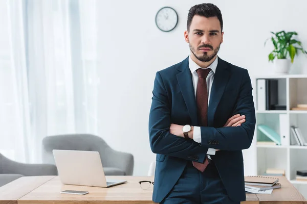 Selbstbewusster junger Geschäftsmann steht mit verschränkten Armen im Büro und blickt in die Kamera — Stockfoto