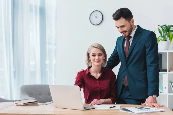 Lächelnde junge Geschäftskollegen, die im Büro gemeinsam auf Laptop schauen — Stockfoto