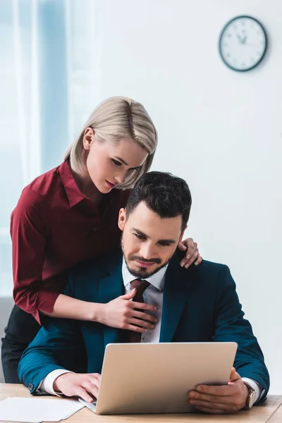 Joven mujer de negocios abrazando apuesto hombre de negocios utilizando el ordenador portátil en la oficina - foto de stock