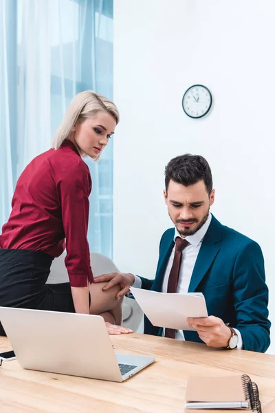 Handsome businessman holding papers and touching knee of young female colleague sitting on table — Stock Photo