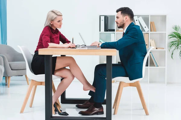 Side view of young couple of business people looking at each other while working together and flirting under table in office — Stock Photo