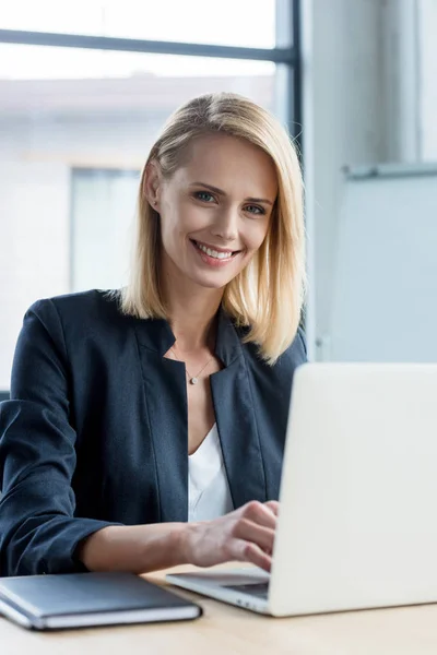 Hermosa mujer de negocios rubia utilizando el ordenador portátil y sonriendo a la cámara - foto de stock