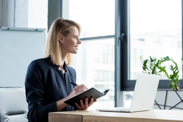 Seitenansicht einer lächelnden Geschäftsfrau, die in Notizbuch schreibt und im Büro mit Laptop arbeitet — Stockfoto