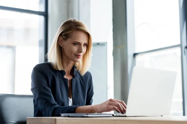 Улыбающаяся профессиональная деловая женщина, работающая с ноутбуком в офисе — стоковое фото