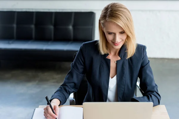 Hochauflösende Ansicht einer lächelnden Geschäftsfrau, die Notizen macht und im Büro mit Laptop arbeitet — Stockfoto