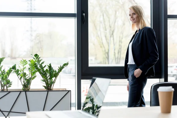 Bela sorridente loira empresária de pé com a mão no bolso e olhando para longe no escritório — Fotografia de Stock