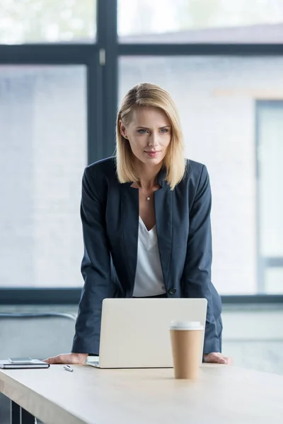 Atractiva mujer de negocios rubia apoyada en la mesa con el ordenador portátil en la oficina - foto de stock