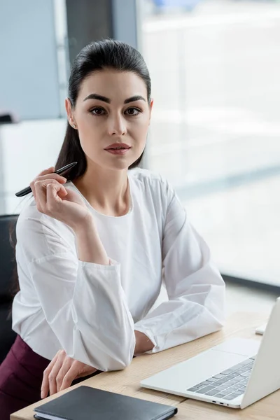 Beautiful young businesswoman holding pen and looking at camera in office — Stock Photo