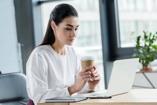 Jeune femme d'affaires tenant tasse en papier et utilisant un ordinateur portable sur le lieu de travail — Photo de stock