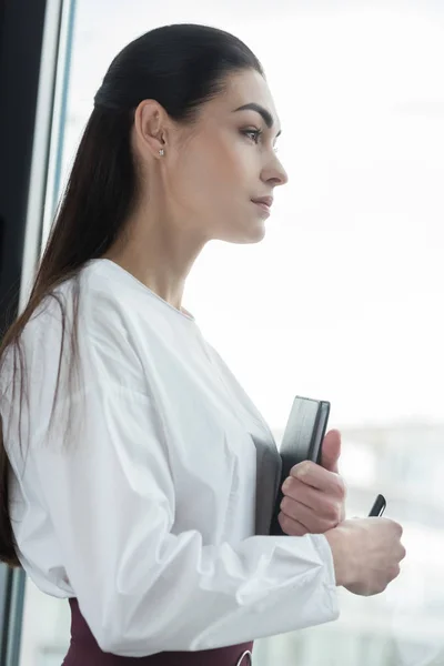 Vista lateral de la joven empresaria pensativa sosteniendo el cuaderno y mirando hacia otro lado - foto de stock