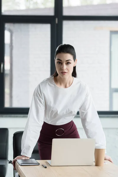 Beautiful young businesswoman looking at camera while leaning at table in office — Stock Photo