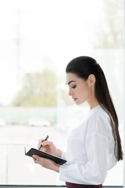 Vue latérale de belle jeune femme d'affaires prenant des notes dans le bloc-notes — Photo de stock