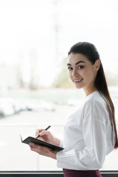 Bela jovem empresária escrevendo em notebook e sorrindo para a câmera — Fotografia de Stock