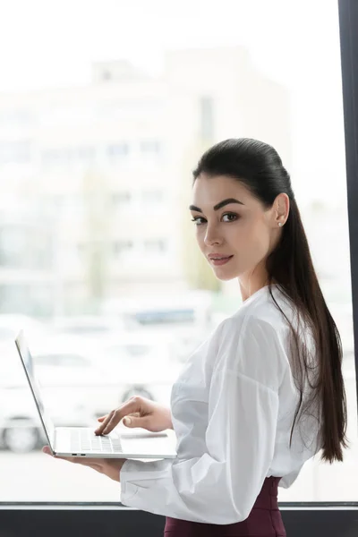 Bela jovem empresária usando laptop e sorrindo para a câmera — Fotografia de Stock