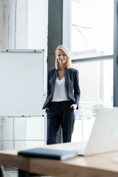 Geschäftsfrau steht mit Händen in den Taschen und lächelt im Büro in die Kamera — Stockfoto