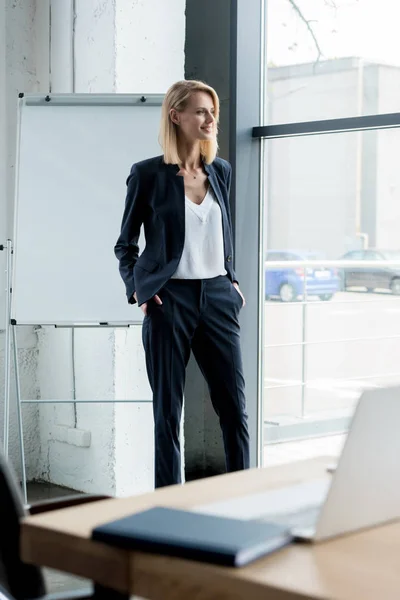 Élégante femme d'affaires blonde debout avec les mains dans les poches et regardant la fenêtre — Photo de stock