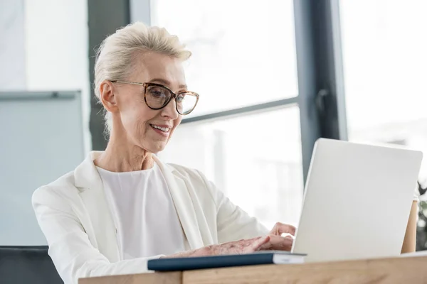 Smiling senior businesswoman in eyeglasses working with laptop in office — Stock Photo