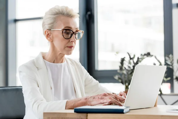 Senior businesswoman in eyeglasses using laptop in office — Stock Photo