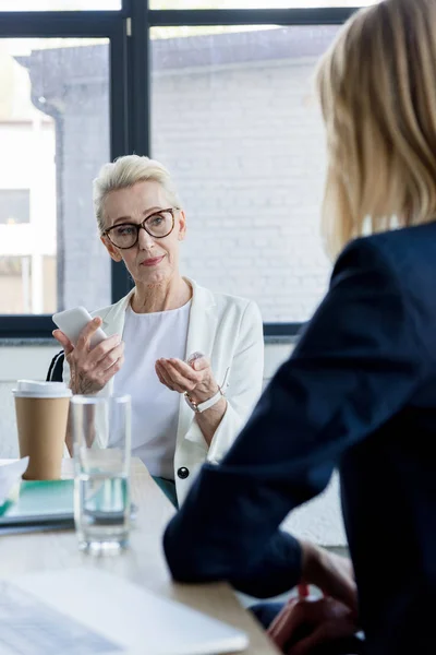 Beautiful businesswoman talking at meeting in office and holding smartphone — Stock Photo