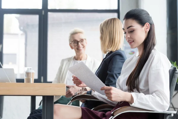Боковой вид красивых деловых женщин, читающих документы и разговаривающих на встрече в офисе — стоковое фото