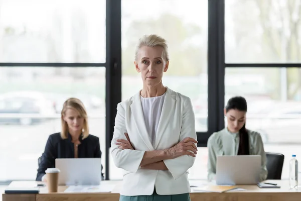 Senior beautiful businesswoman standing with crossed arms and looking at camera in office — Stock Photo