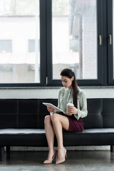 Attrayant femme d'affaires en utilisant une tablette et en tenant du café pour aller sur le canapé dans le bureau — Photo de stock