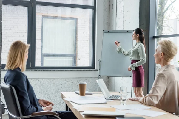 Femme d'affaires écrit sur un tableau à feuilles mobiles et présente un projet à ses collègues lors d'une réunion au bureau — Photo de stock