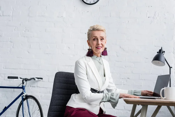 Donna d'affari anziana sorridente usando il computer portatile in ufficio moderno — Foto stock