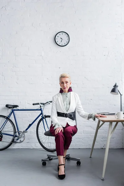 Elegante Seniorin arbeitet in modernem Büro mit Fahrrad — Stockfoto