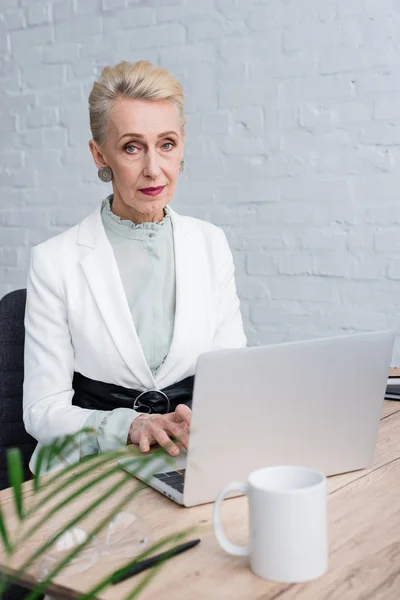 Donna d'affari anziana caucasica che utilizza il computer portatile sul posto di lavoro con tazza di caffè — Foto stock