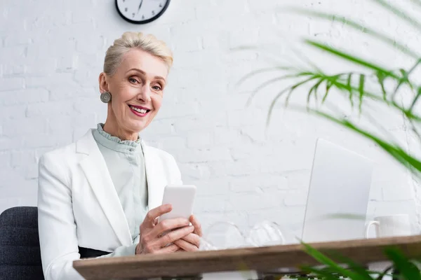 Glückliche Seniorin nutzt Smartphone am Arbeitsplatz mit Laptop — Stockfoto