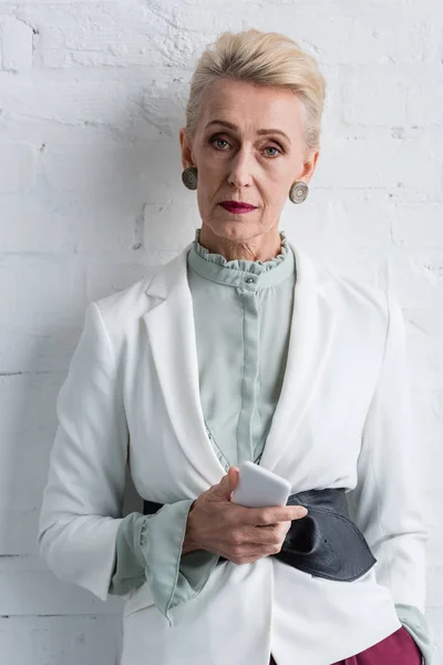 Красивая элегантная старшая деловая женщина со смартфоном, позирующим у белой стены — стоковое фото