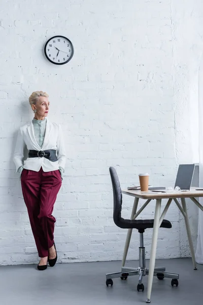 Elegante donna d'affari anziana in posa vicino al posto di lavoro con laptop e caffè — Foto stock
