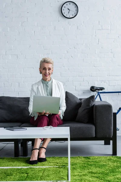 Smiling senior business woman using laptop on sofa in office with bike — Stock Photo