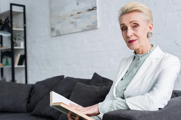 Елегантна старша жінка читає книгу і сидить на дивані — стокове фото