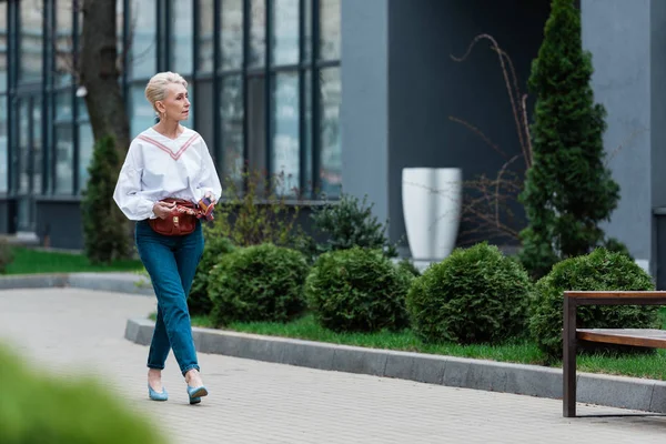 Mujer mayor en traje de moda con bolsa de cintura caminando en el parque - foto de stock