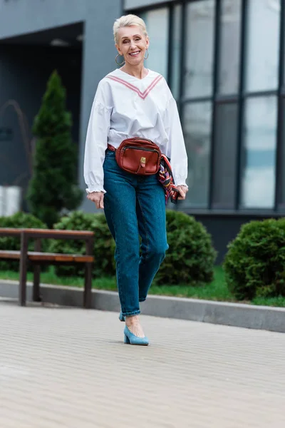 Femme âgée souriante en tenue tendance avec sac de taille marchant dans le parc — Photo de stock