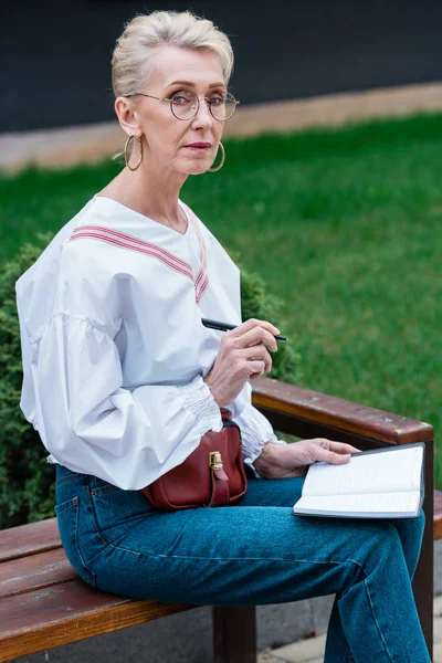 Стильна старша жінка, що пише в щоденнику, сидячи на лавці в парку — стокове фото