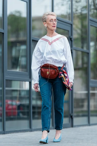 Mujer mayor en traje de moda escuchando música con auriculares mientras camina en la ciudad urbana - foto de stock