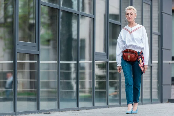 Schöne Seniorin in stylischem Outfit, die mit Kopfhörern Musik hört, während sie in der Stadt spaziert — Stockfoto