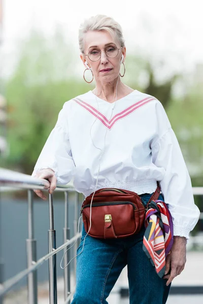 Senior woman listening music with earphones and standing near railings — Stock Photo