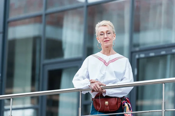 Femme âgée en tenue tendance écouter de la musique avec des écouteurs et debout près des balustrades dans la ville urbaine — Photo de stock