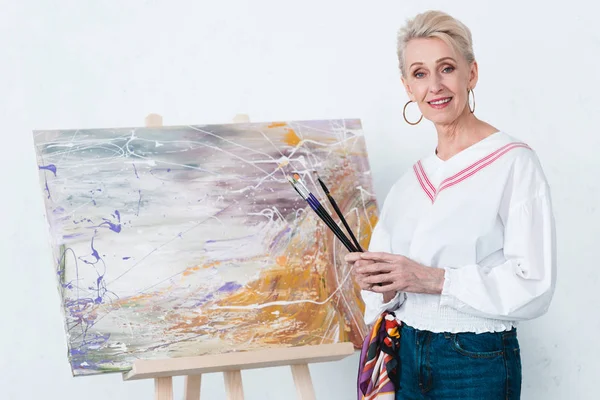 Senior elegant female artist holding paintbrushes and standing near painting on easel in workshop — Stock Photo