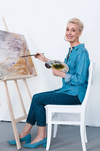 Senior woman painting on easel with paintbrush and palette in workshop — Stock Photo