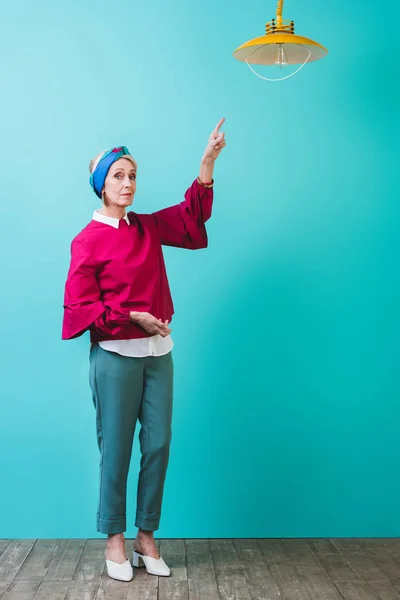 Mujer mayor de moda apuntando a la lámpara en la habitación con la pared de color turquesa - foto de stock