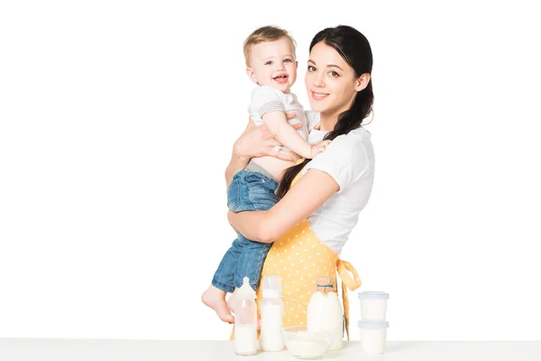 Young mother in apron holding baby boy at table with children food isolated on white background — Stock Photo