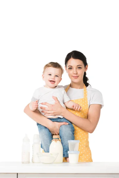 Mãe jovem com filho em mãos na mesa com garrafa de leite, recipientes de plástico, tigela e mamadeiras isoladas em fundo branco — Fotografia de Stock